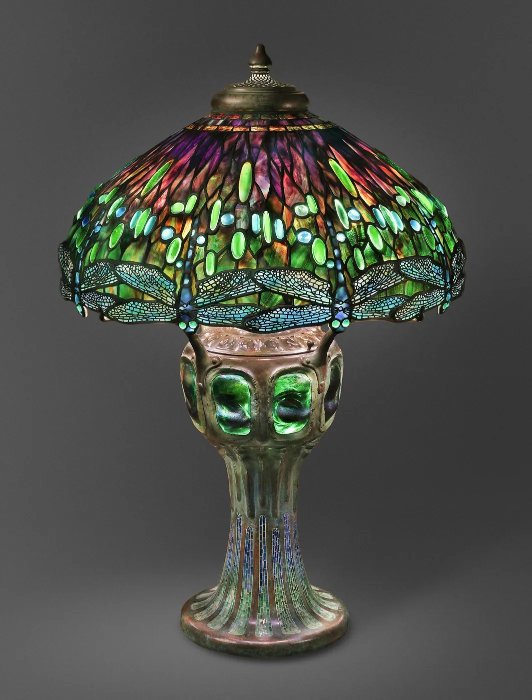Leaded-glass table lamp with a domed shade of green, blue and violet glass ringed by dragonflies, on a bronze base set with green glass on grey