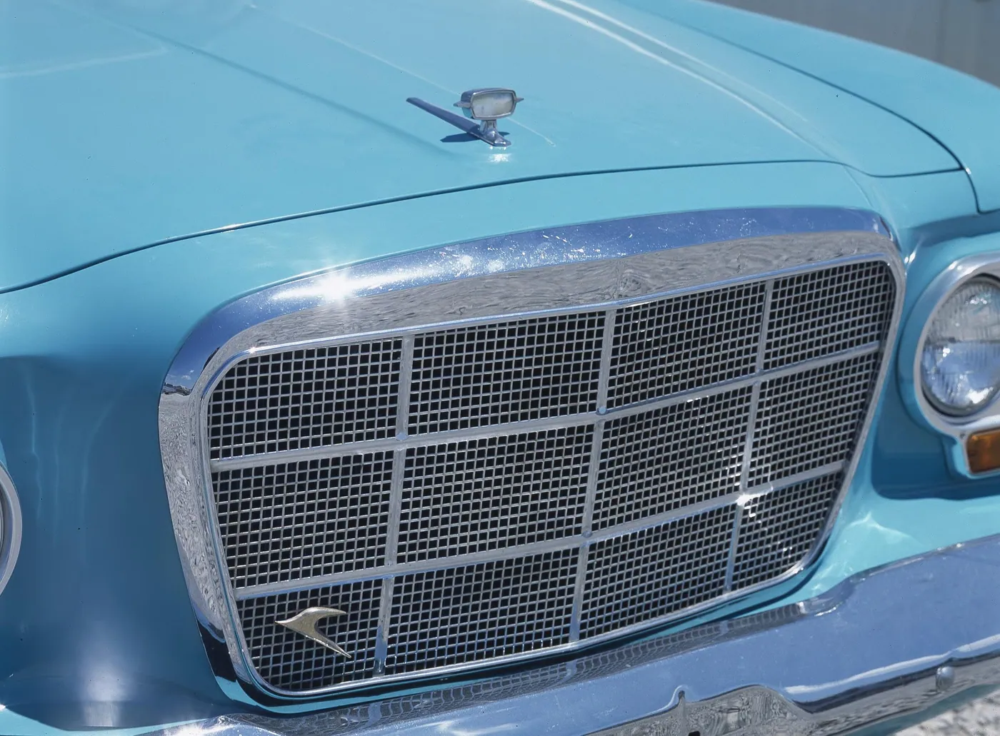 Close view of a turquoise vintage car's chrome-framed grille, round headlight and hood ornament gleaming in bright sunlight.
