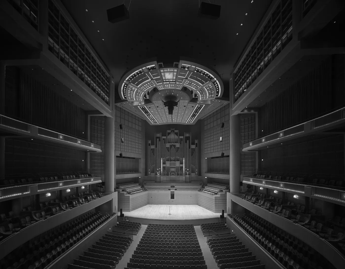 Black-and-white view of an empty concert hall with tiered balconies, a lit stage and a pipe organ beneath a suspended canopy.