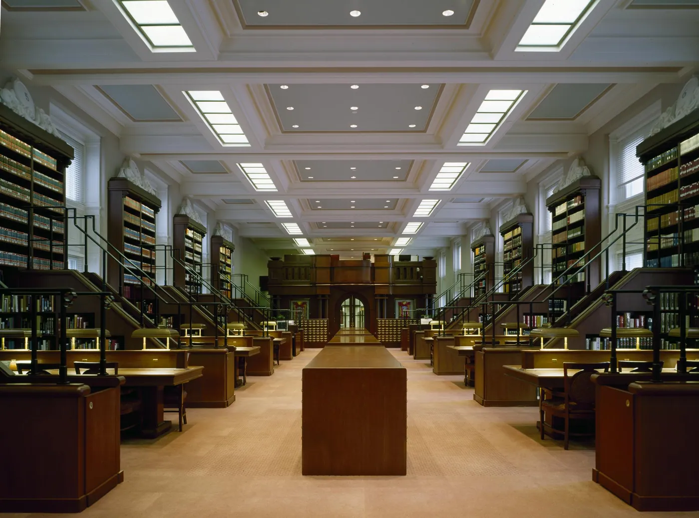 Long, symmetrical library reading room with coffered skylit ceiling, two tiers of book stacks and rows of wooden reading desks with brass lamps