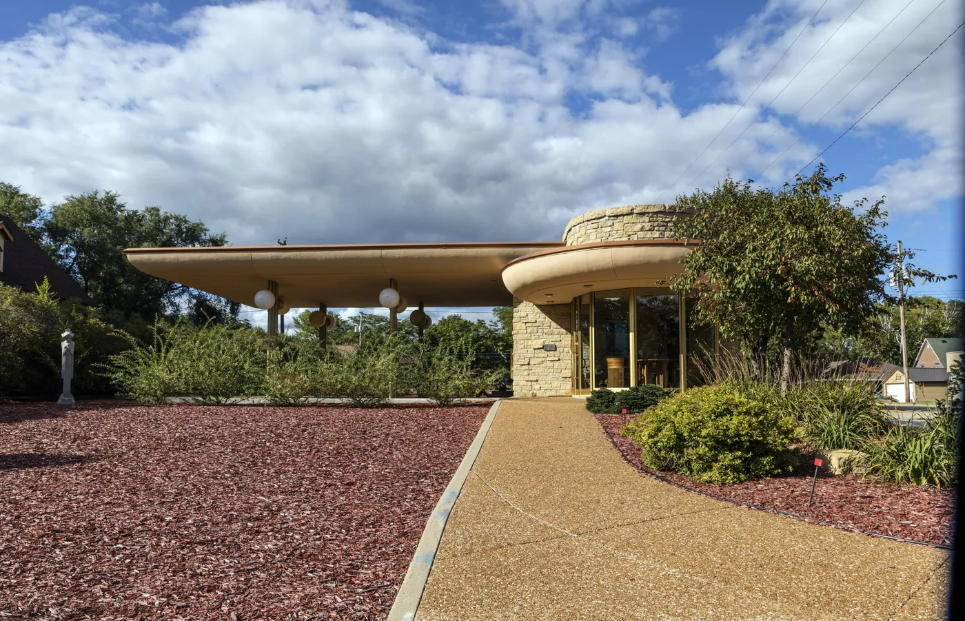 A low modernist bank of honey-coloured stone with a long cantilevered roof and a curved glass entrance, reached by a pale path through planting