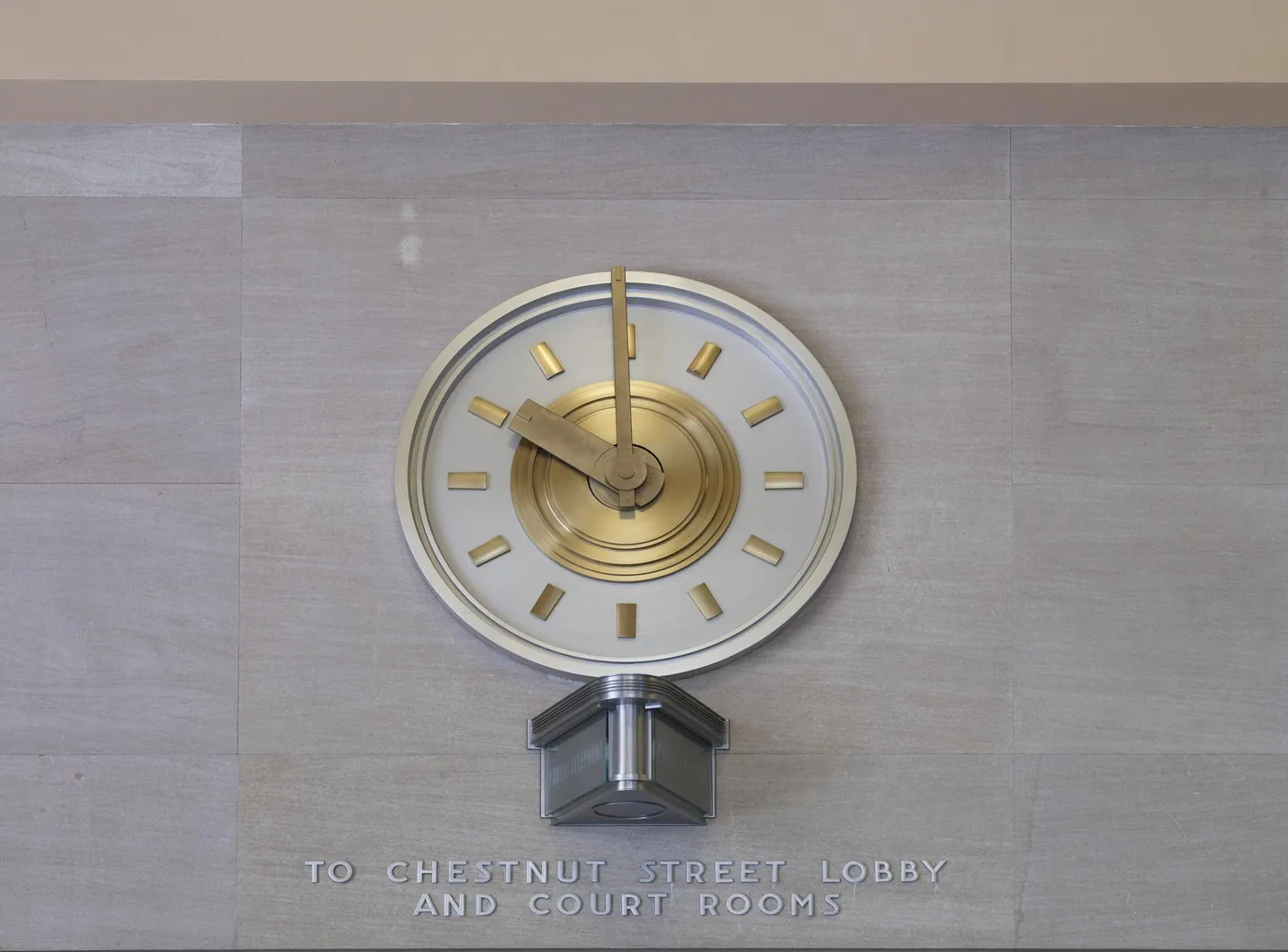 A round Moderne wall clock with gold hands and hour bars on pale stone, above carved lettering pointing to the lobby and court rooms