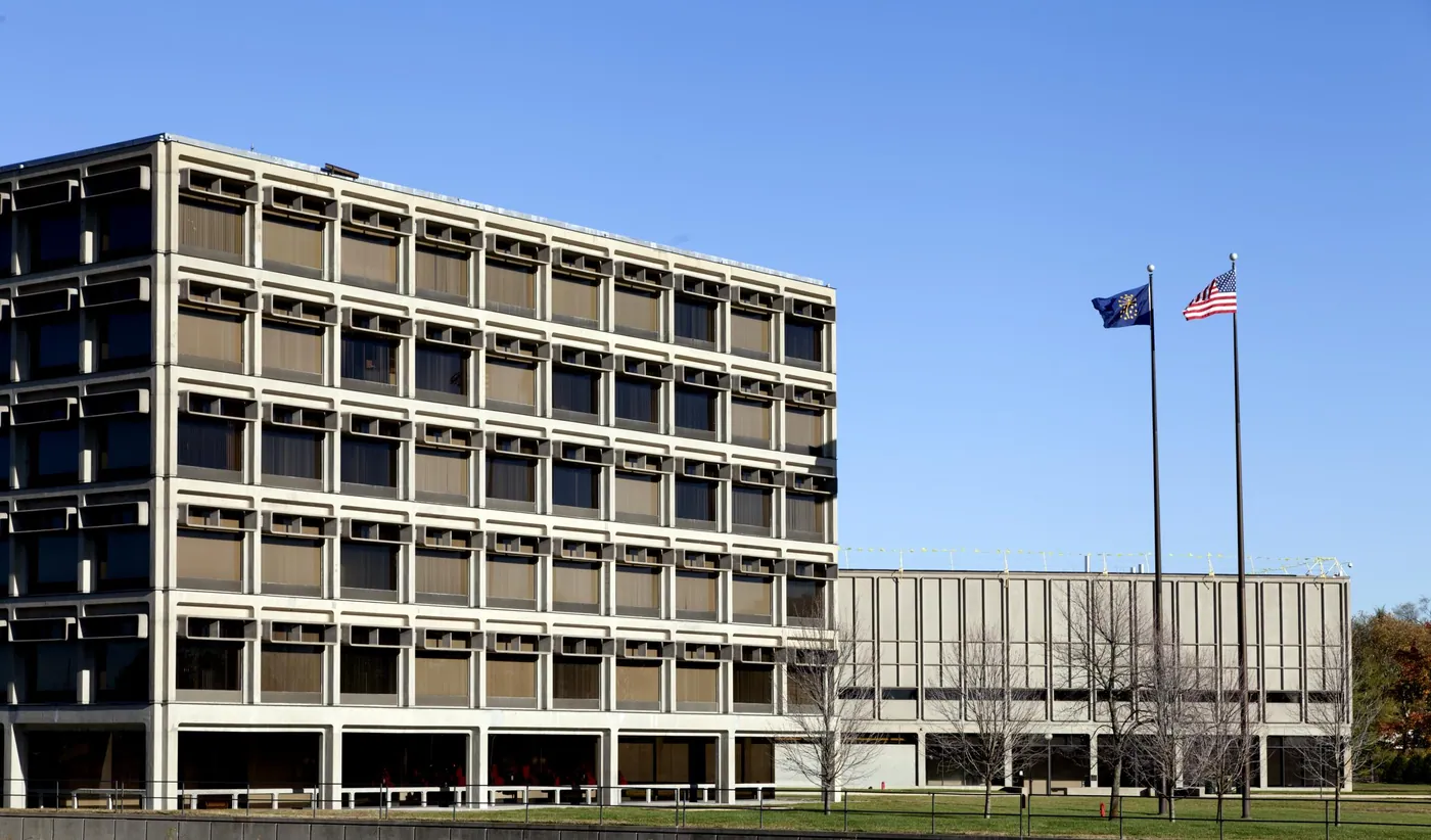 A mid-century office building with a deep gridded concrete facade and shaded windows beside a low wing, two flags flying against a clear blue sky