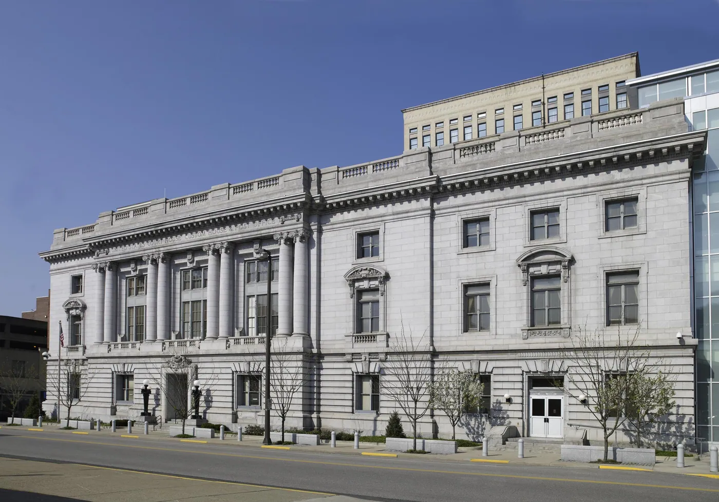 Long pale granite Beaux-Arts courthouse with a columned upper storey and balustraded roofline under a clear blue sky, young trees along the street