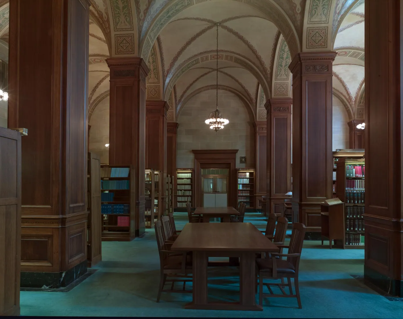 Hushed law library with vaulted painted ceilings, dark wood piers, bookshelves, a hanging chandelier and a long wooden reading table with chairs