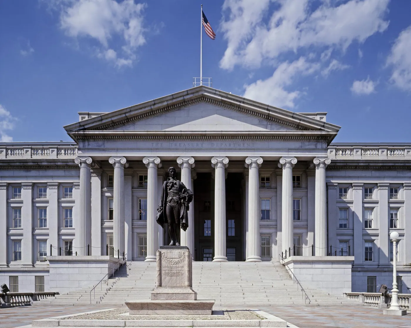 Columned south portico of the U.S. Treasury Building under a blue sky with clouds, a bronze statue on a pedestal before the broad steps