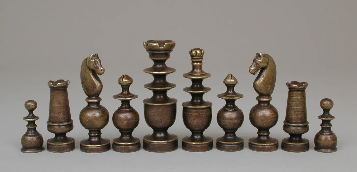 A row of antique bronze chessmen, from pawns and rooks to knights, bishops, queen and king, standing side by side on grey