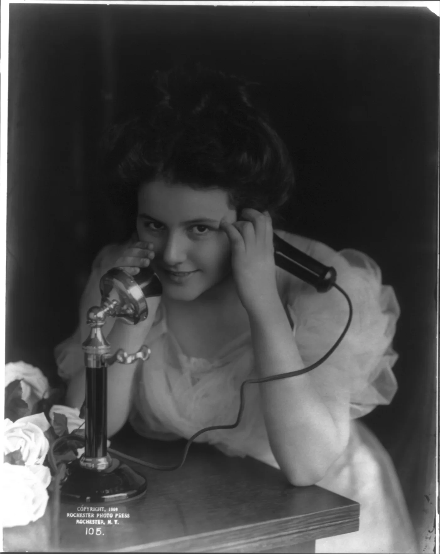 A young woman speaks into the transmitter of an upright telephone around 1909