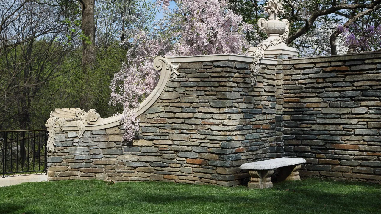 Curving stone garden wall with a carved scroll and urn, pink blossoming trees above, and a stone bench on a green lawn