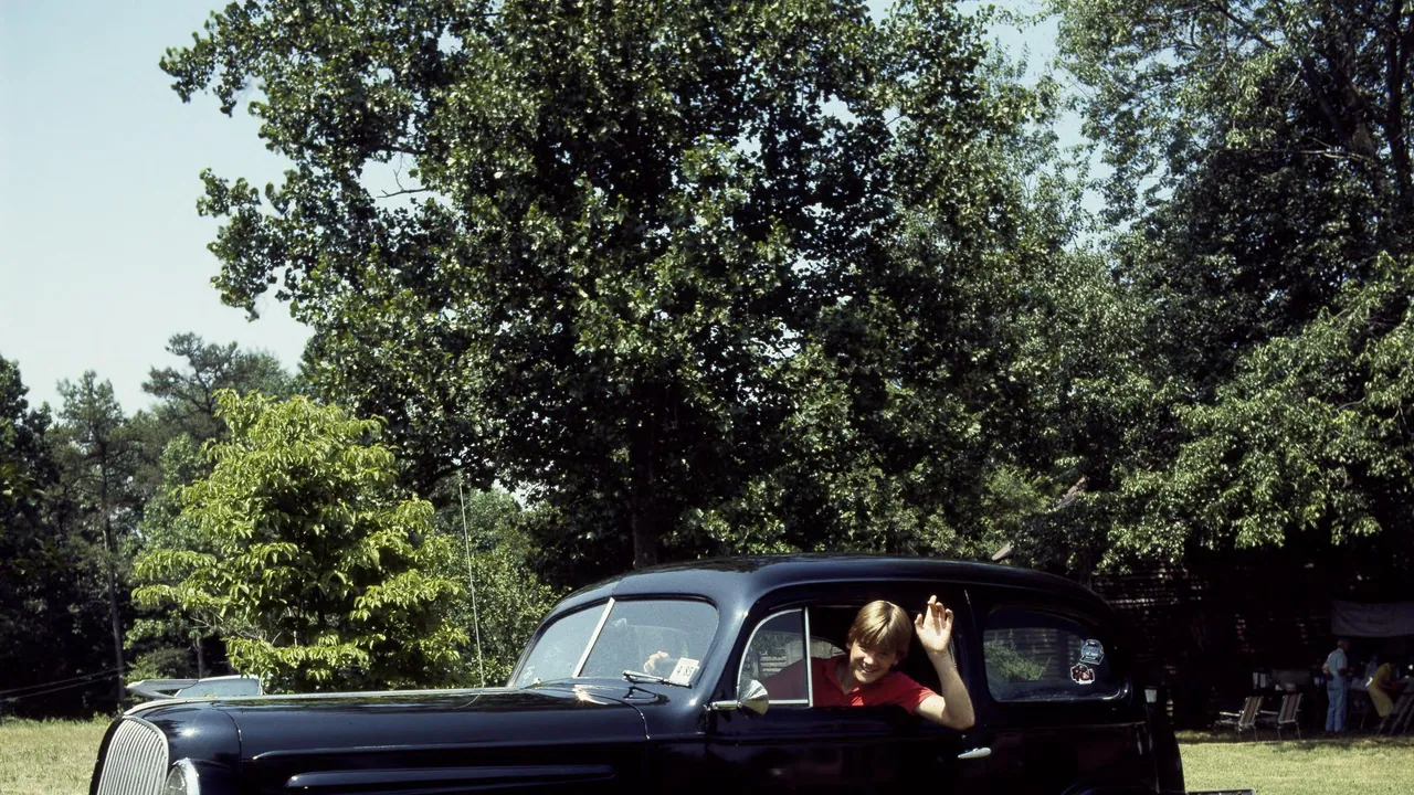 A driver waves from the open window of a black vintage car parked on a green lawn beneath mature trees